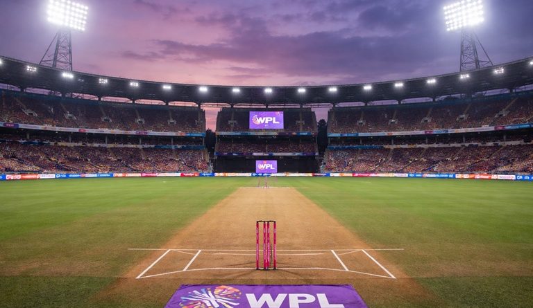 A wide-angle view of DY Patil Stadium in Navi Mumbai during a Women’s Premier League night match, showing the empty cricket pitch under floodlights with WPL branding and sunset sky