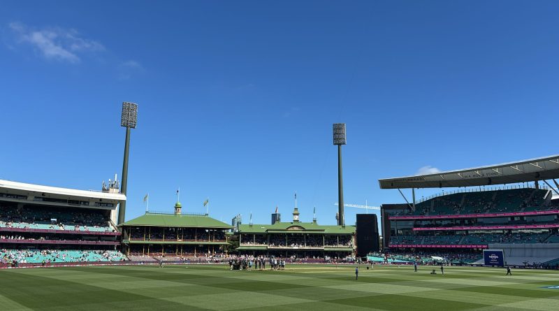 Wide view of the Sydney Cricket Ground on day four of the fifth Ashes Test, with players gathered near the pitch under clear blue skies.