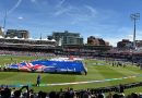 Grand Atmosphere at Lord’s: England vs Australia Women’s T20 World Cup Final! 🏏✨ A wide, eye-level shot taken from the crowded spectator stands looking out over the sunlit outfield of Lord's Cricket Ground during a pre-match ceremony. In the center of the lush green field, a large group of people holds up a massive Australian flag flat against the grass, while a second large flag is being unrolled further back. Spectators in the foreground are holding up smartphones to capture the scene under a bright blue sky with light, wispy clouds. The historic grandstands and a tall stadium floodlight tower are visible in the background under the clear afternoon light.