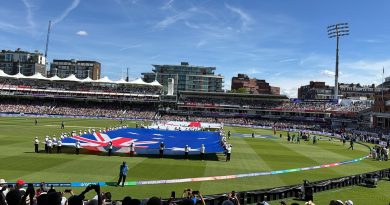 A wide, eye-level shot taken from the crowded spectator stands looking out over the sunlit outfield of Lord's Cricket Ground during a pre-match ceremony. In the center of the lush green field, a large group of people holds up a massive Australian flag flat against the grass, while a second large flag is being unrolled further back. Spectators in the foreground are holding up smartphones to capture the scene under a bright blue sky with light, wispy clouds. The historic grandstands and a tall stadium floodlight tower are visible in the background under the clear afternoon light.
